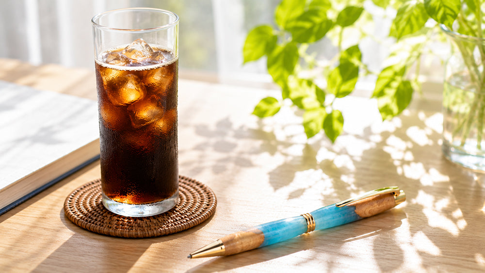 夏の明るい光が差し込むデスク。アイスコーヒーの横に、透き通る水色やパステルブルーのハイブリッドウッドペンが涼しげに置かれている写真。